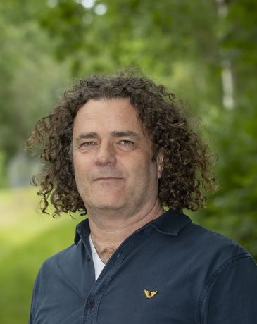 Ludger Sluys  met krullend haar, gekleed in een lichtblauw shirt met patroon, lacht tegen een effen beige muur.