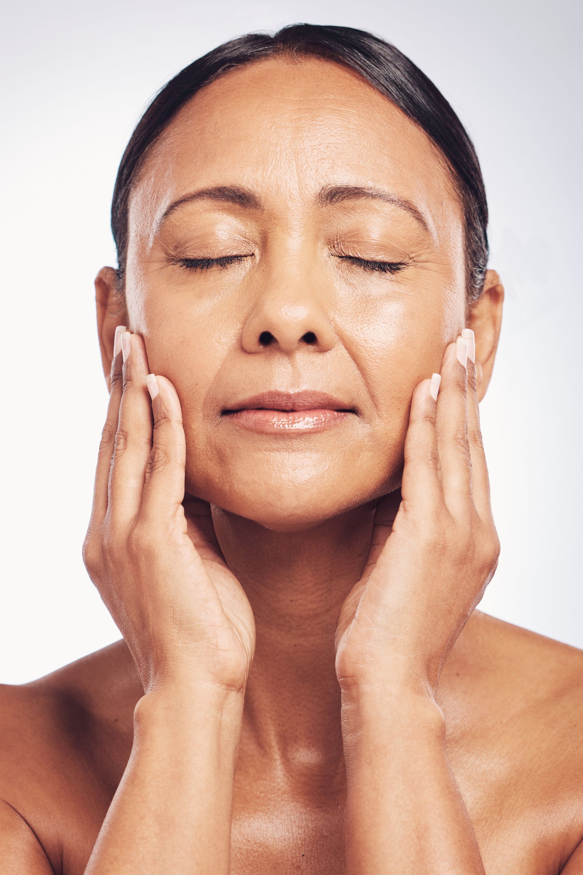 Woman with closed eyes, hands on cheeks, skin glowing. Light background.