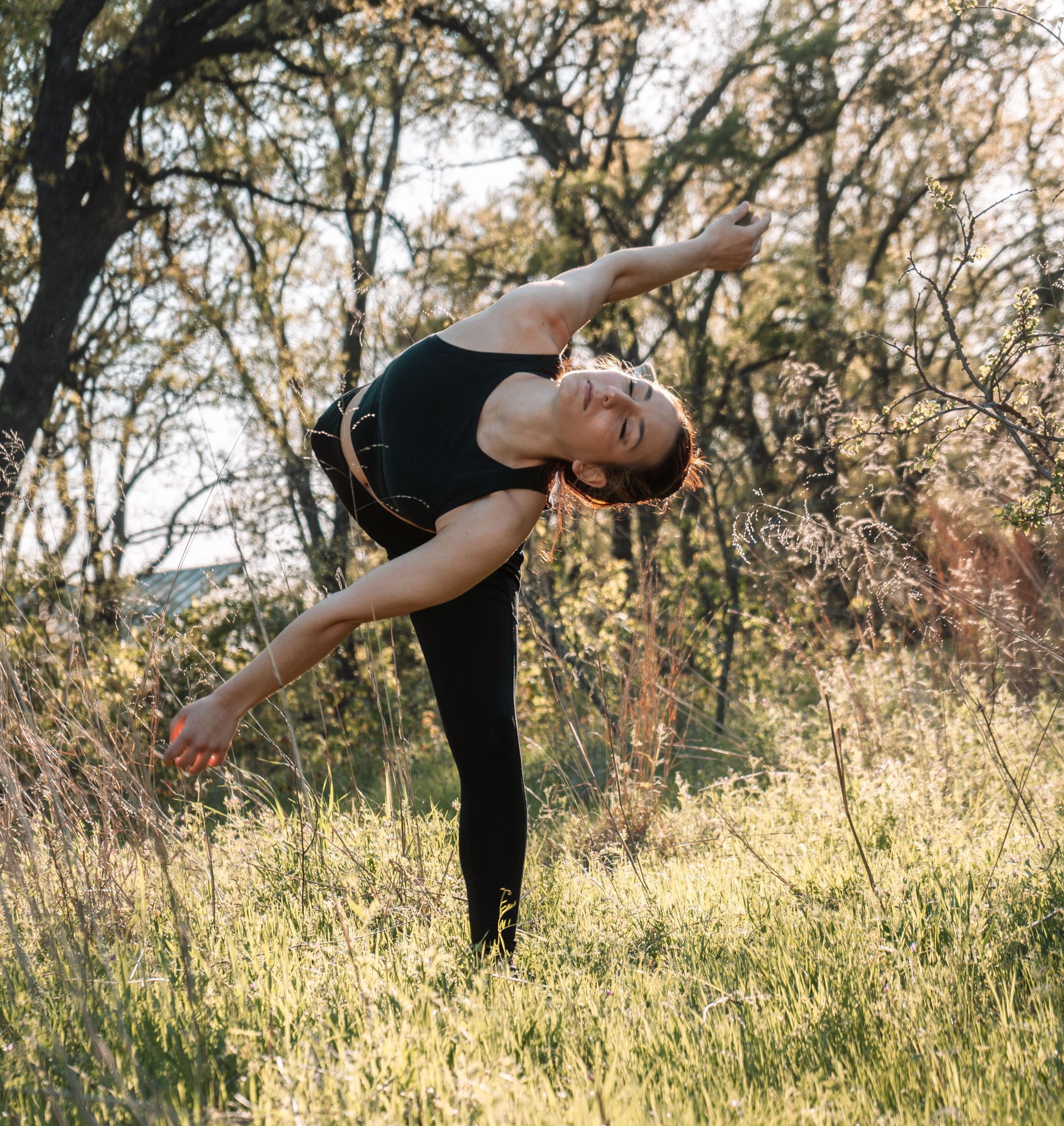Woman in black activewear doing a balancing yoga pose in a grassy field with trees.