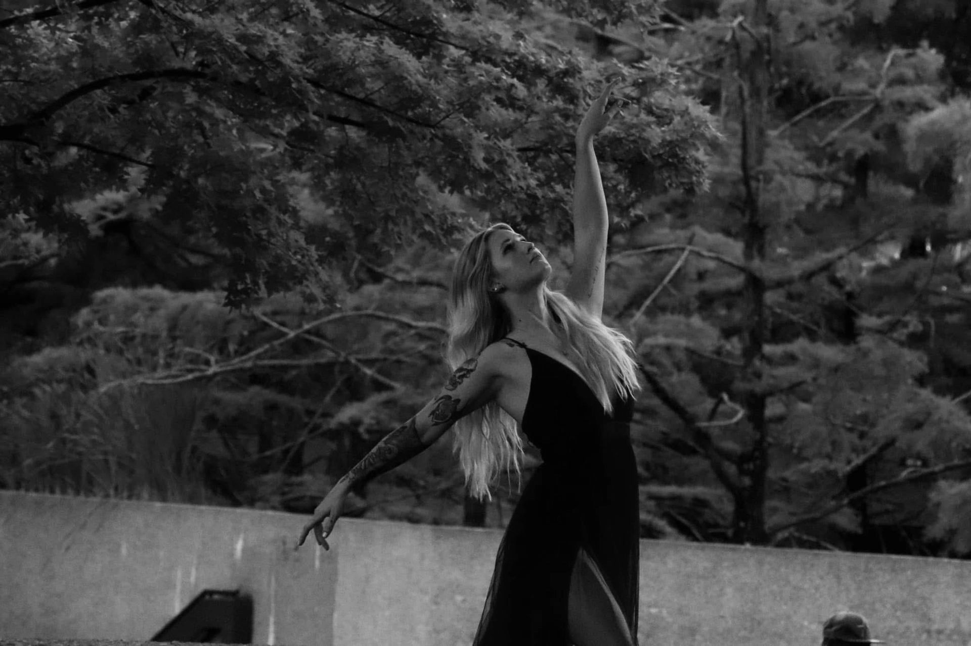 Woman in a black dress dances gracefully outdoors, arms raised toward the sky. Black and white photo.