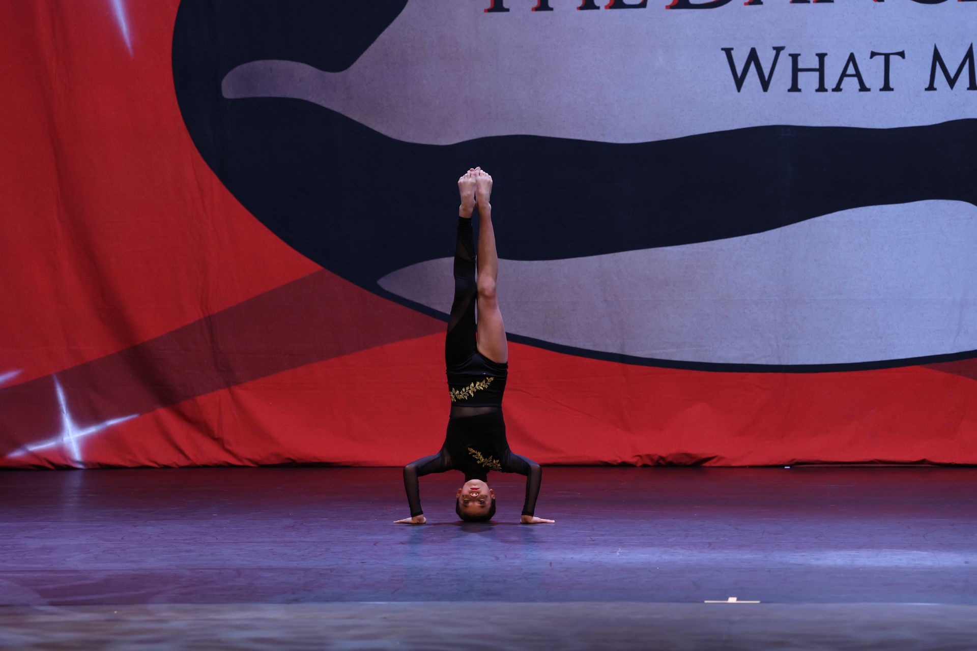 A person in a black leotard performs a handstand on a stage, legs extended, against a red and blue backdrop.
