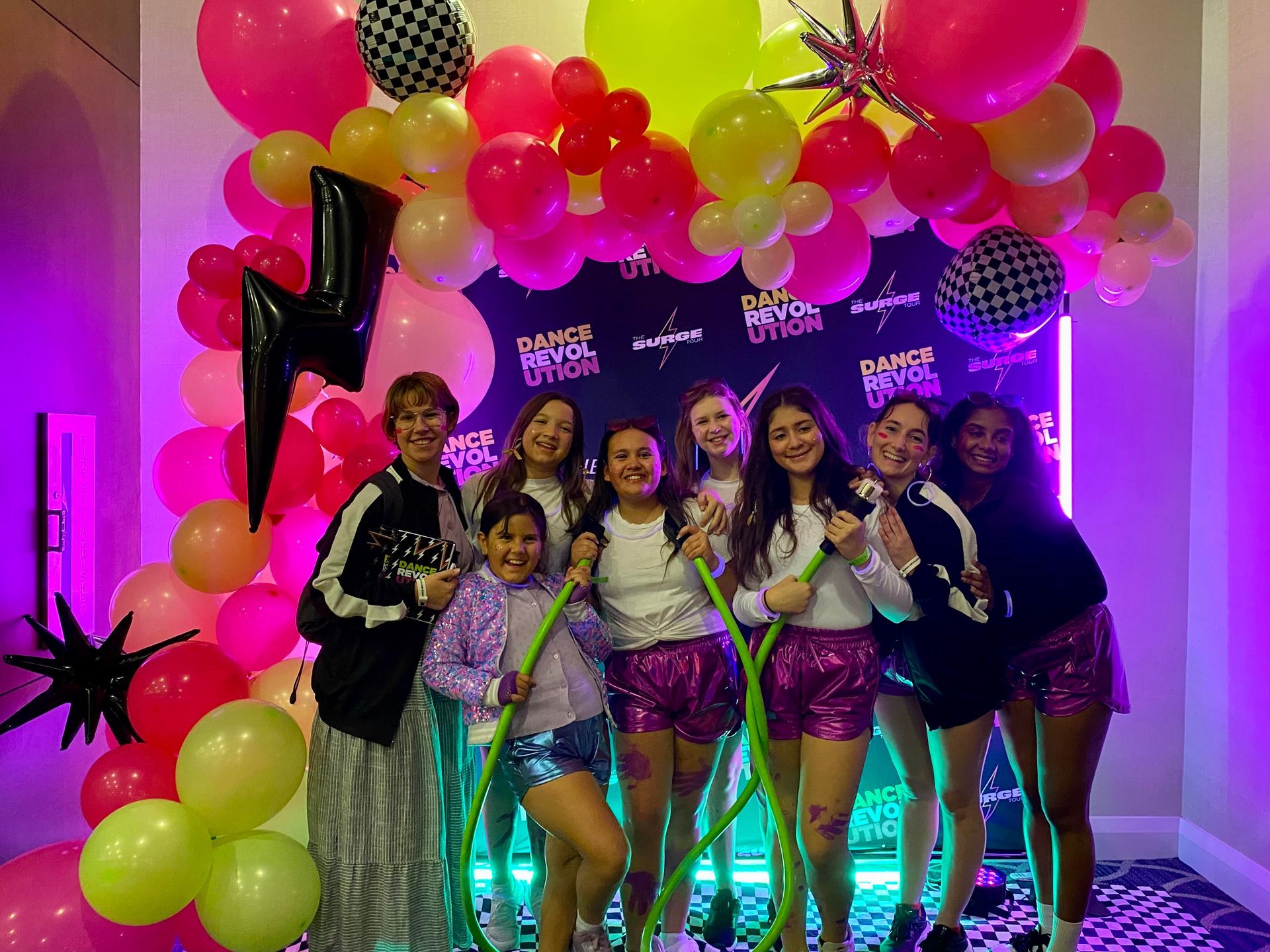 Group of people posing in front of a colorful balloon arch. They are wearing metallic shorts and holding props.