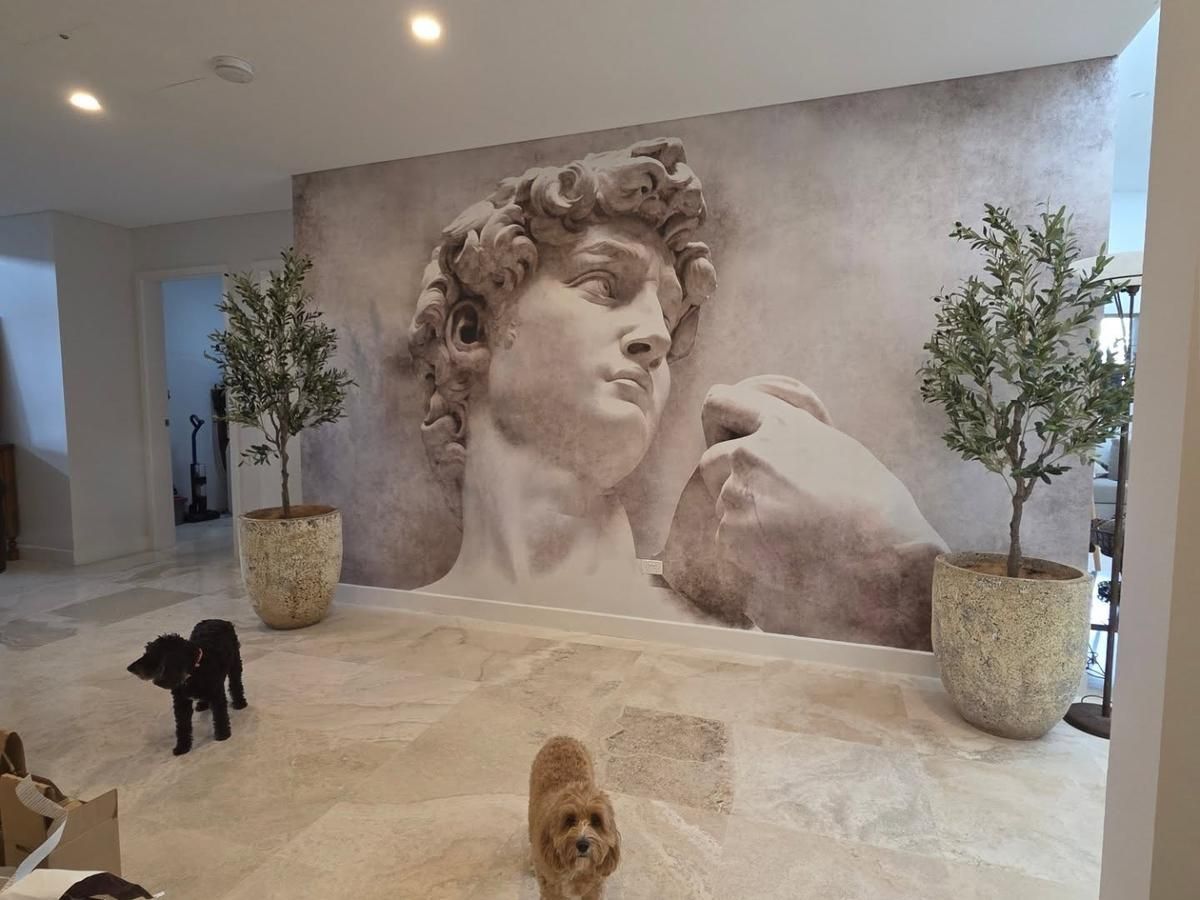 Mural wallpaper of Michelangelo's David dominates a large interior wall, flanked by two potted olive trees on a marble floor. Two small dogs, one brown and one black, are visible in the room.