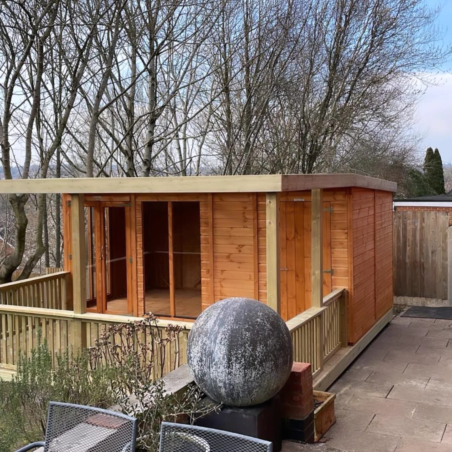 Wooden garden office with deck, brown walls, flat roof, and open doors. A large stone sphere sits nearby.