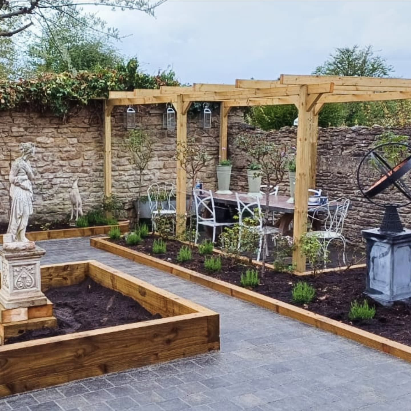 A backyard garden with a wooden pergola over a dining area and stone statue, and stone walls.