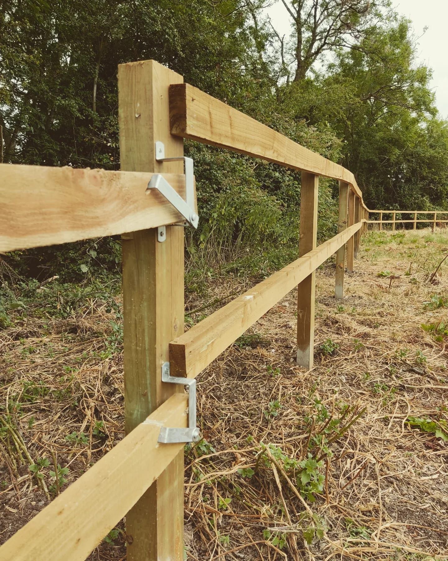 Wooden fence posts with two horizontal rails, secured by metal brackets, in a grassy field.