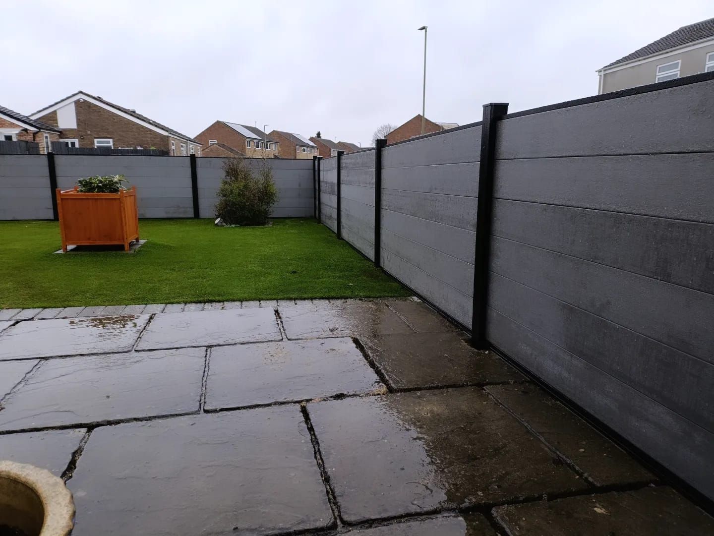Gray composite fence in a backyard on a cloudy day. Stone patio in the foreground, green grass in the middle.