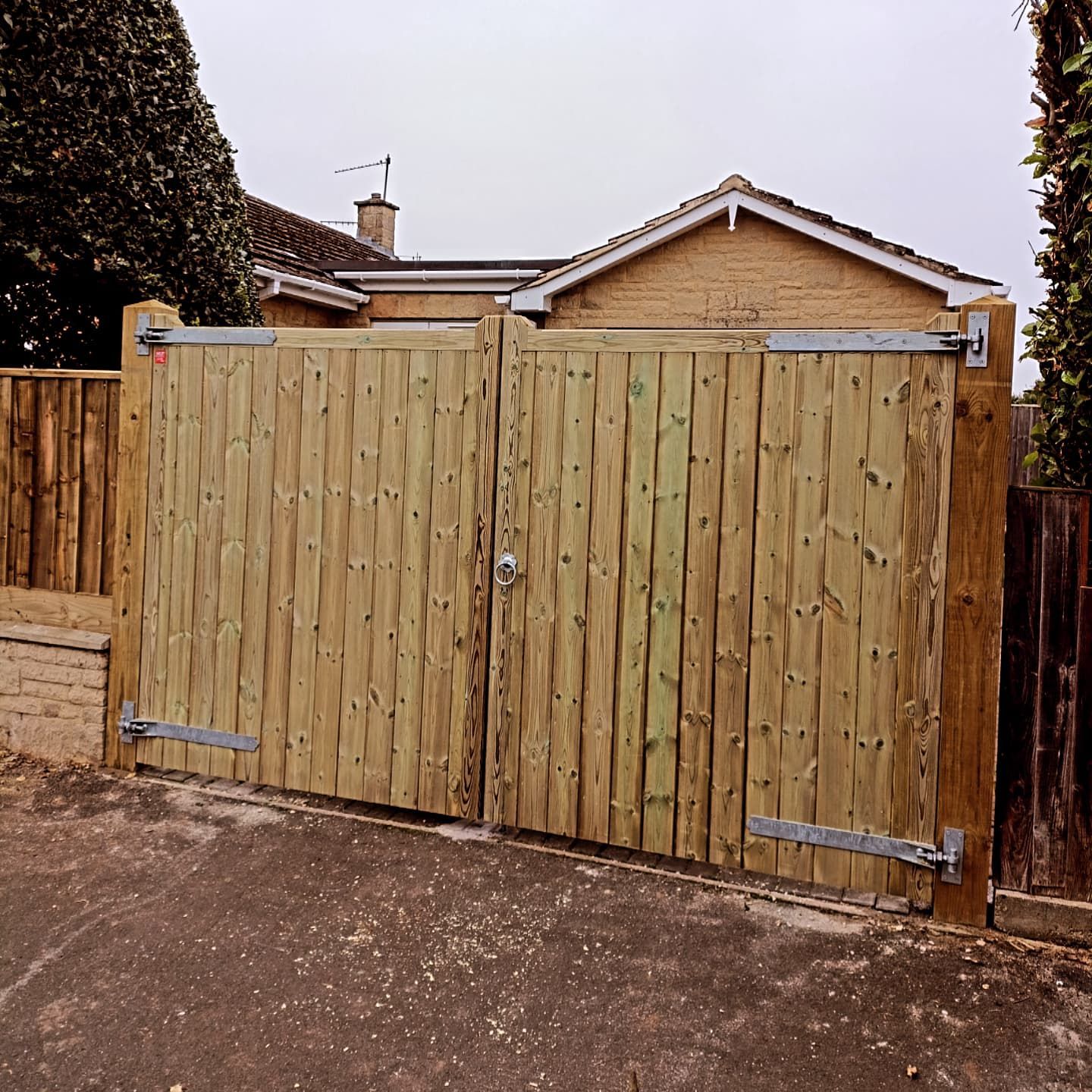 Wooden double gates, light brown with metal hinges, set in a driveway, leading to a house.