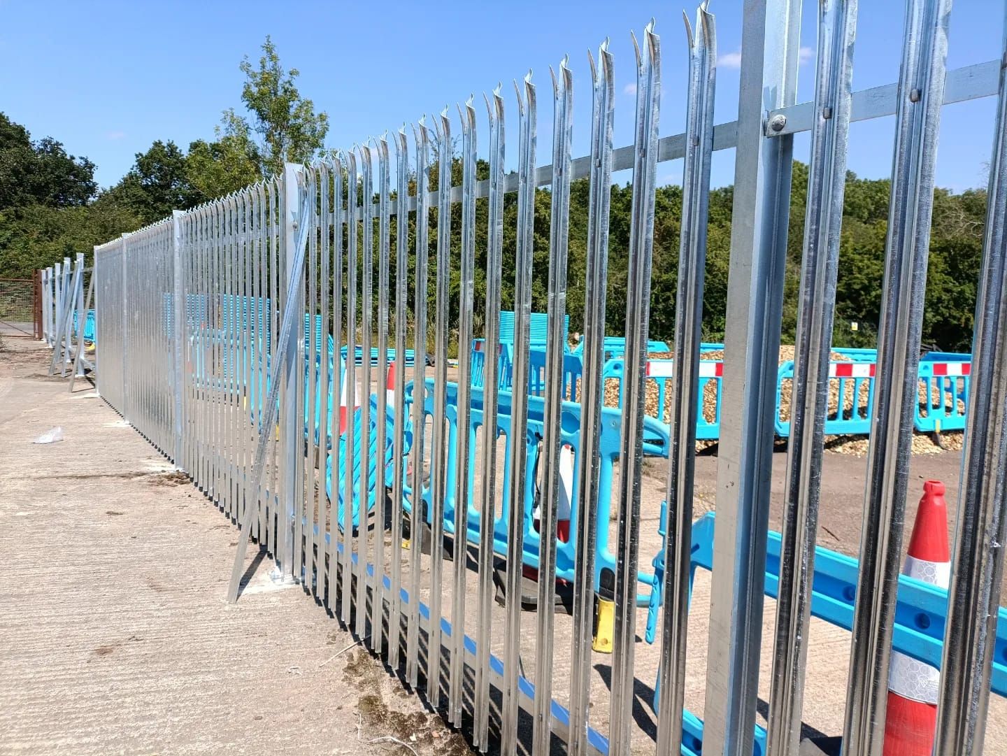 Galvanized metal security fence with sharp points; construction site with blue barriers and trees.