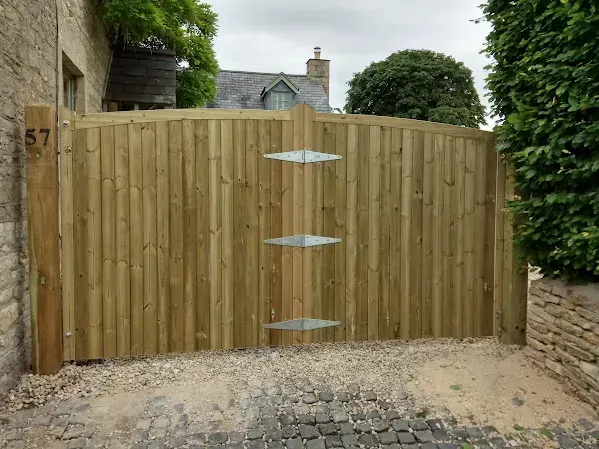Wooden arched gate with three hinges, gravel driveway, building and greenery.