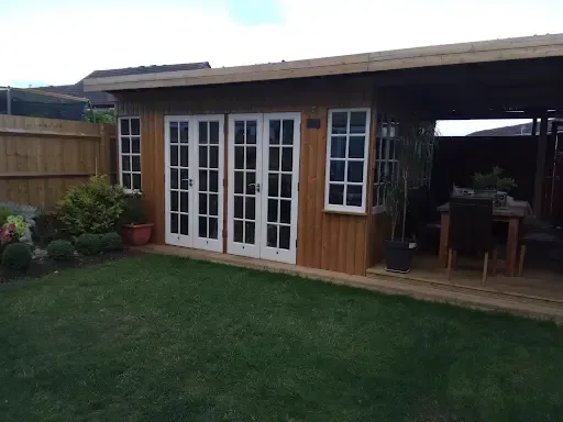 Wooden garden shed with French doors, windows, and an attached covered deck, surrounded by grass and a wooden fence.