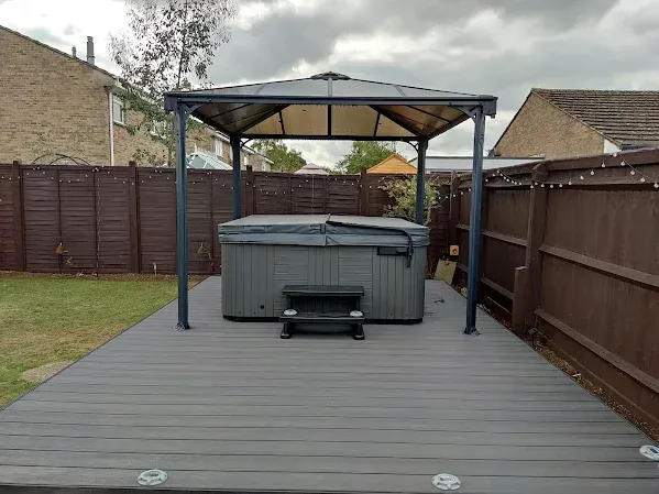 Hot tub on a gray deck under a canopy in a backyard, brown fence, cloudy sky.