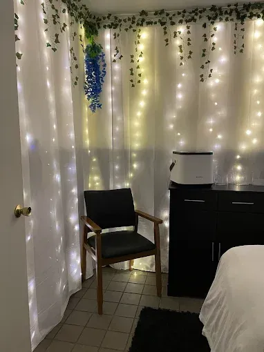 Cozy room with string lights, white curtains, a black chair, and a dresser beside a bed