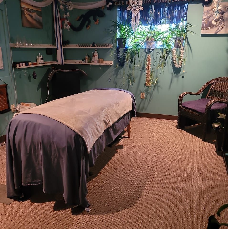 Massage room with teal walls, covered table, woven chair, and plants by a bright window.