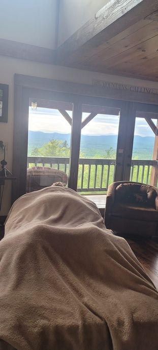 Cozy cabin porch view through glass doors, with mountains and blue sky beyond a wooden railing.