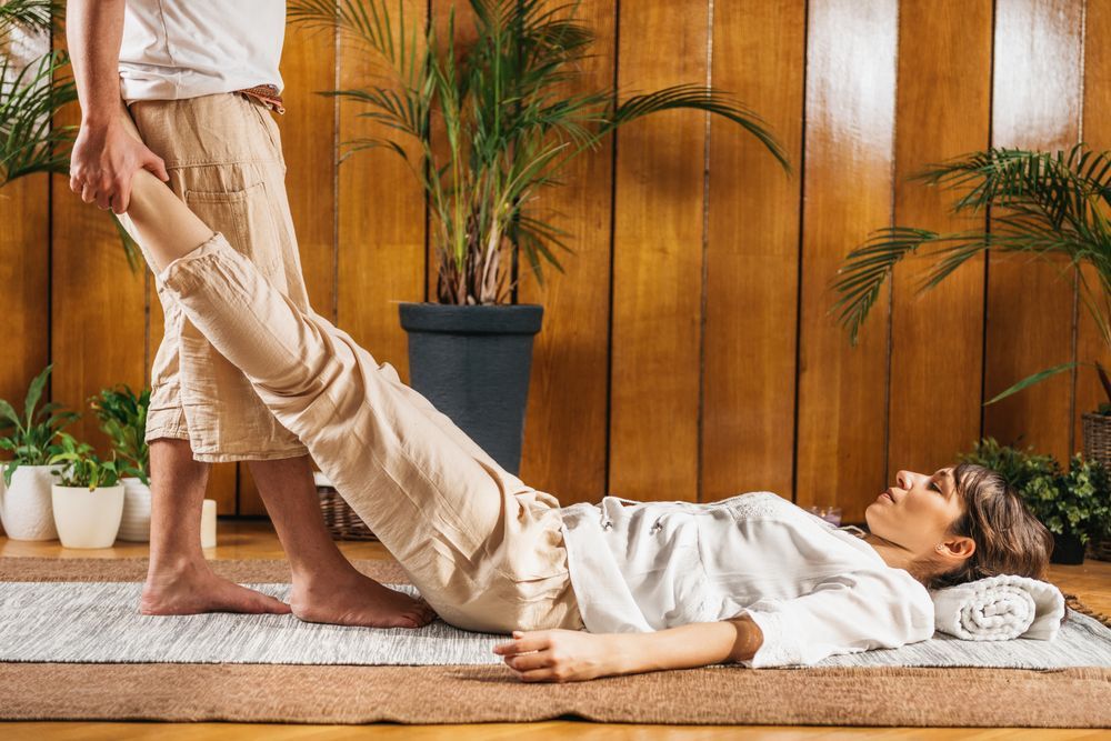 Person assisting another in a yoga stretch on a mat in a wooden room with potted plants