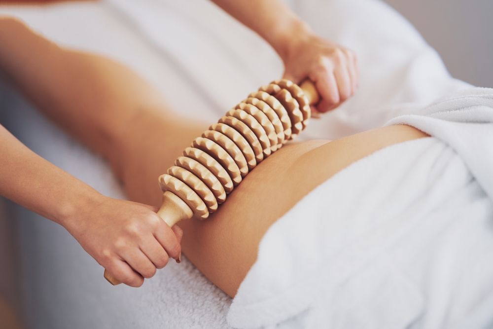 Hands holding a stacked wooden massage roller on a person’s thigh under a white towel