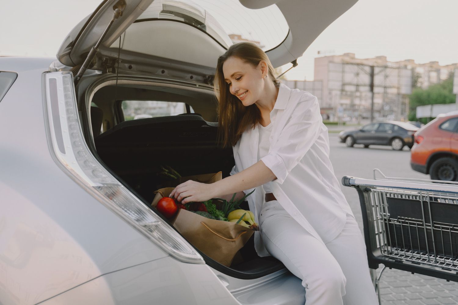 Woman loading groceries into the open trunk of a white car in a parking lot