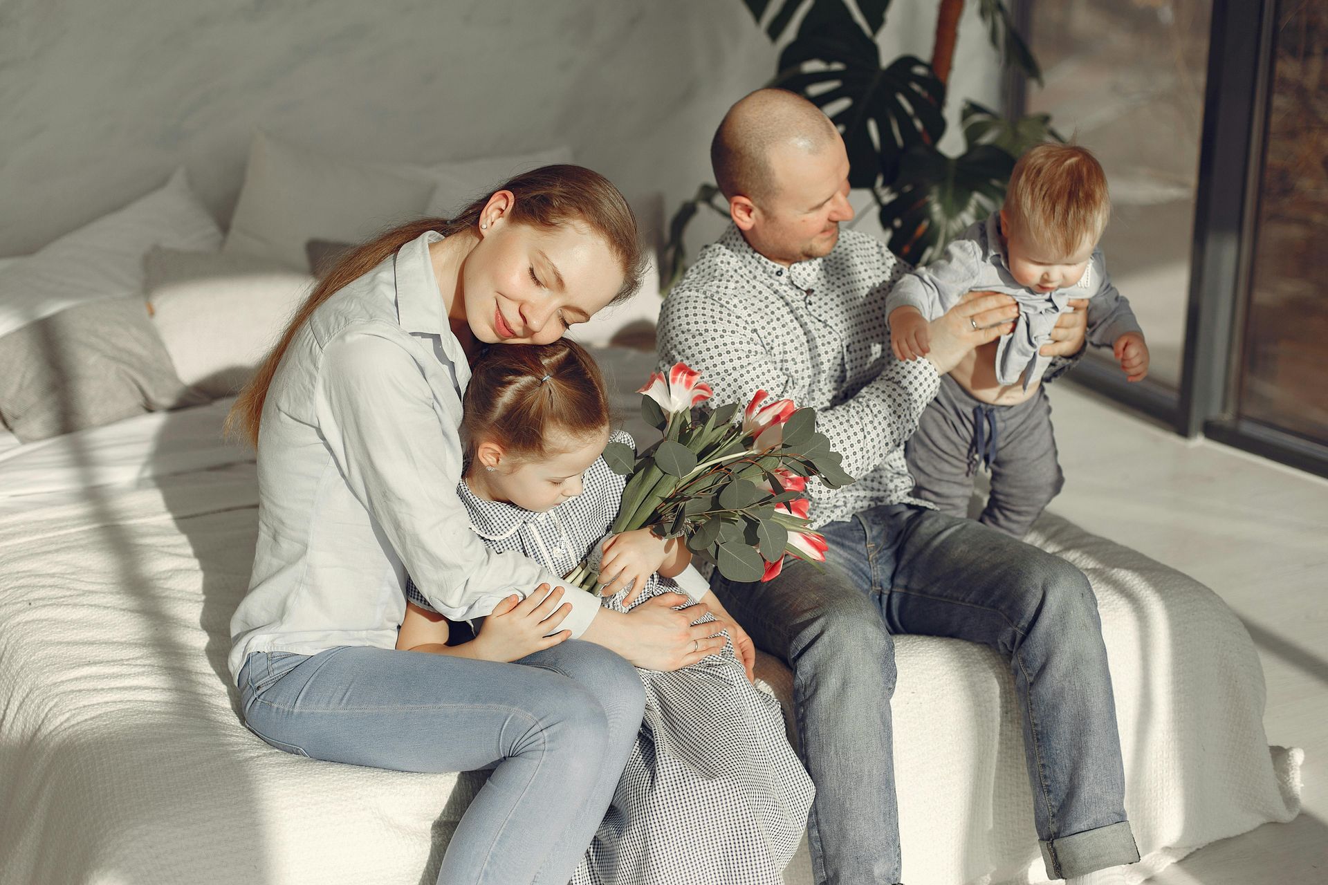 Family sitting on a sunlit couch, cuddling with a bouquet of flowers nearby