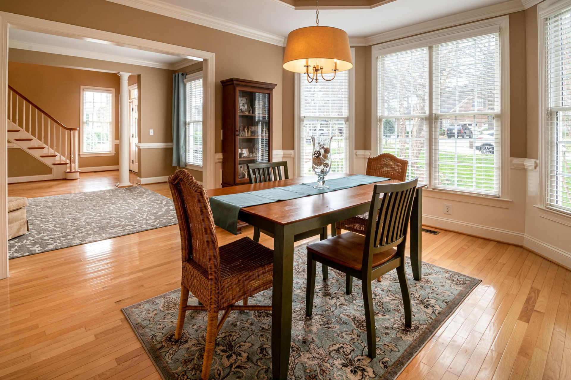 Dining room with wooden table and chairs, bay windows, and a view into a hallway and staircase.