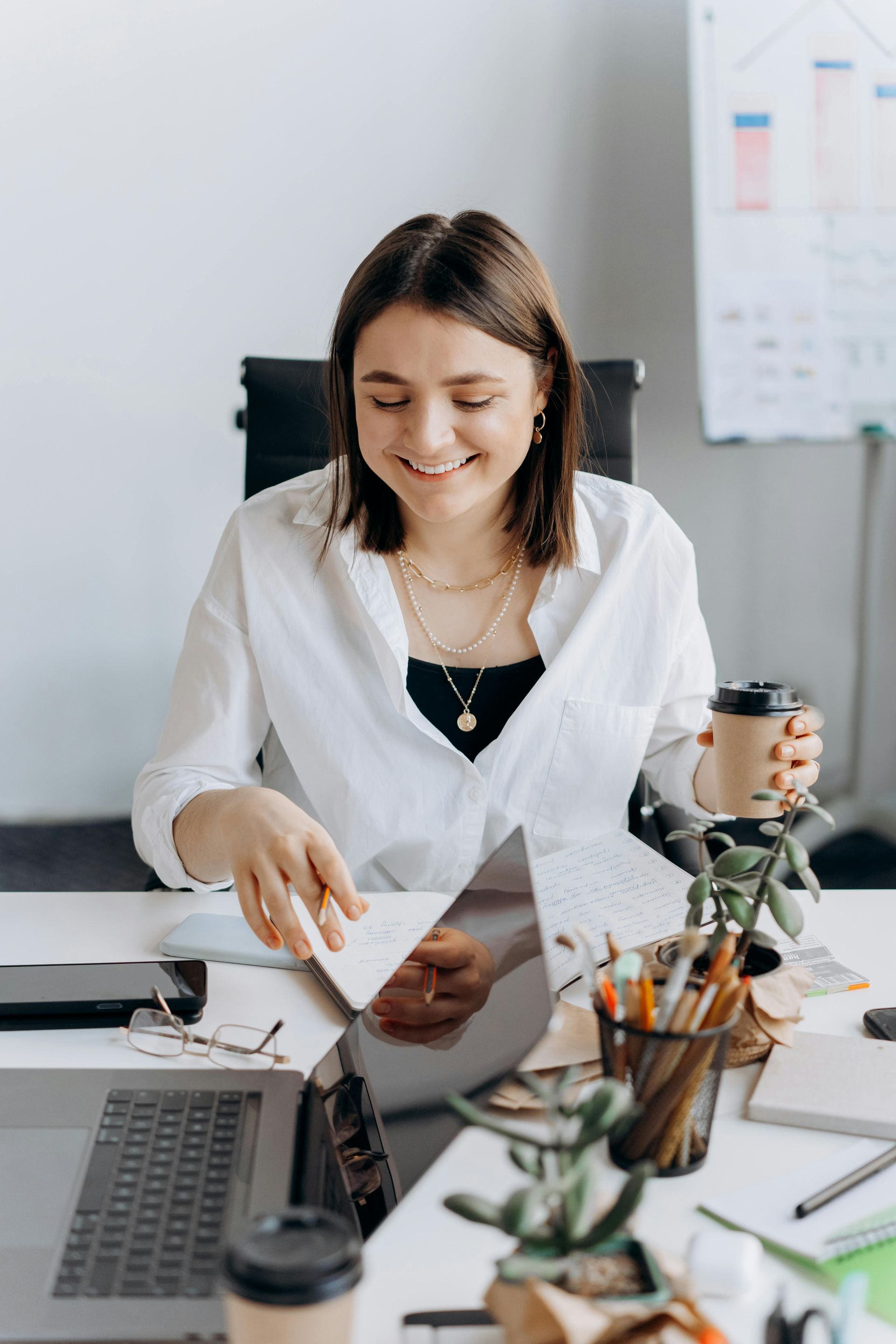 Smiling person reviews documents at a desk with laptop, coffee, and office supplies
