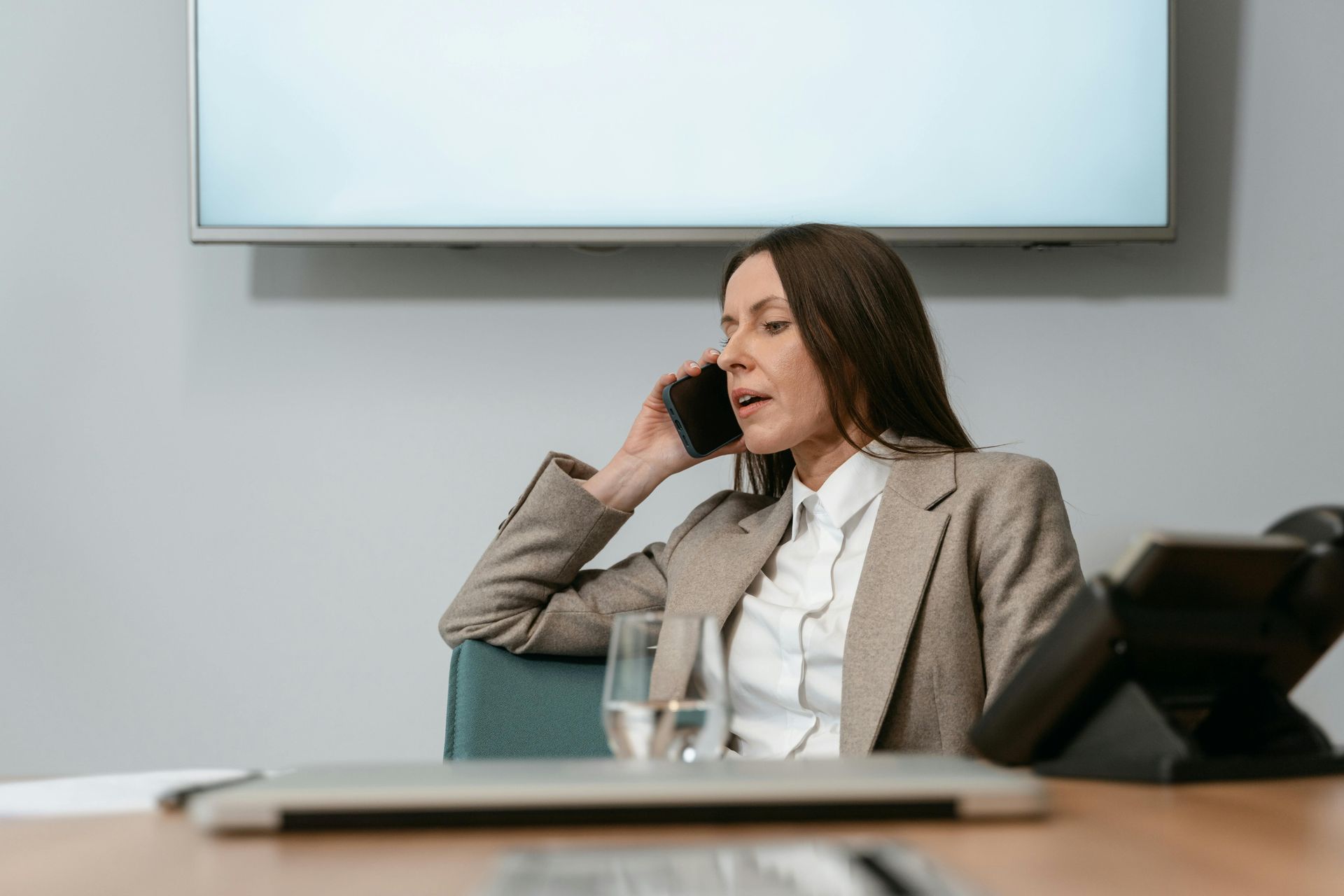 Woman in a beige blazer talks on the phone in a conference room, seated beside a glass of water.