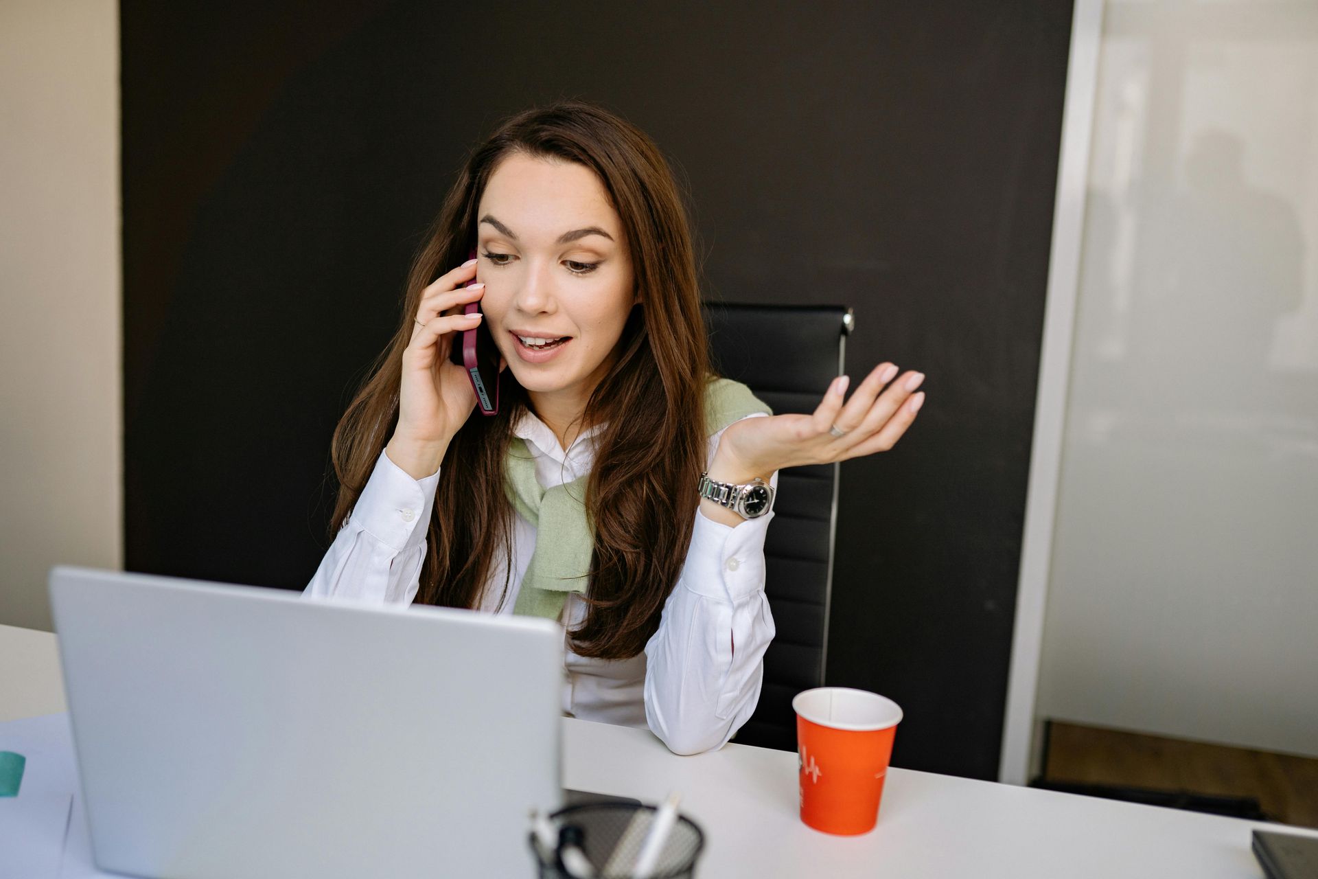 Woman on a phone call at a laptop, gesturing in a modern office with a red cup on the desk.