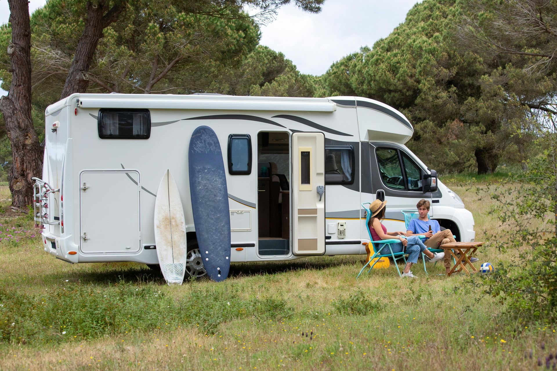 Camper van parked in a grassy field with surfboards outside and two people relaxing at a picnic table.