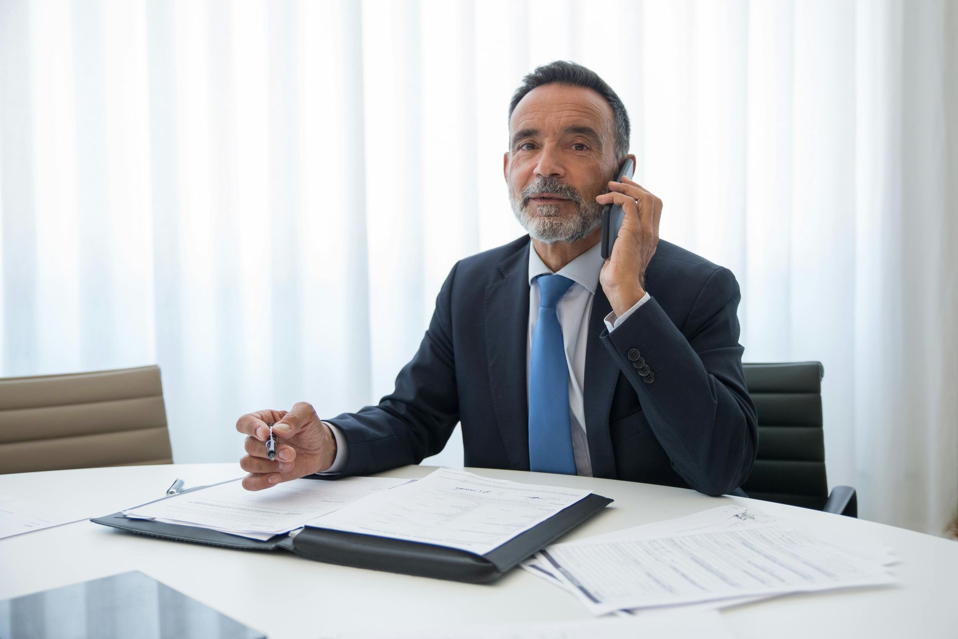 Professional in a suit speaking on a phone at a bright office desk with open documents and a laptop