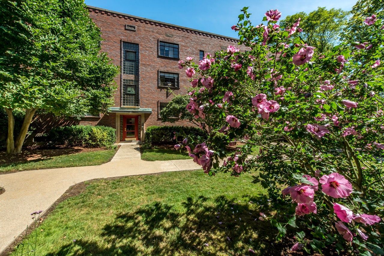 Brick apartment building entrance with sidewalk and pink flowering bushes in front.