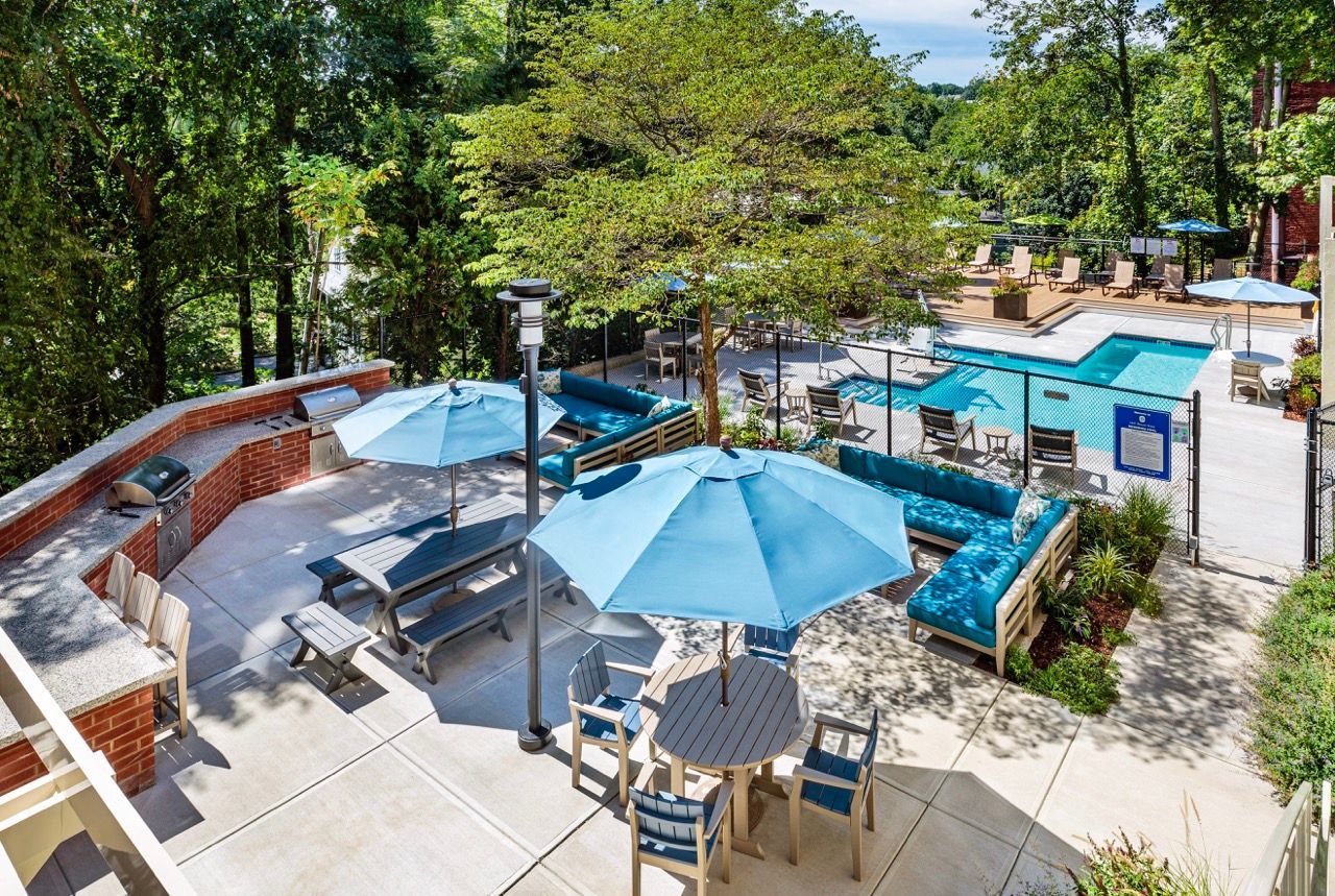 Outdoor pool deck in a tree-lined apartment community with blue seating and umbrellas.