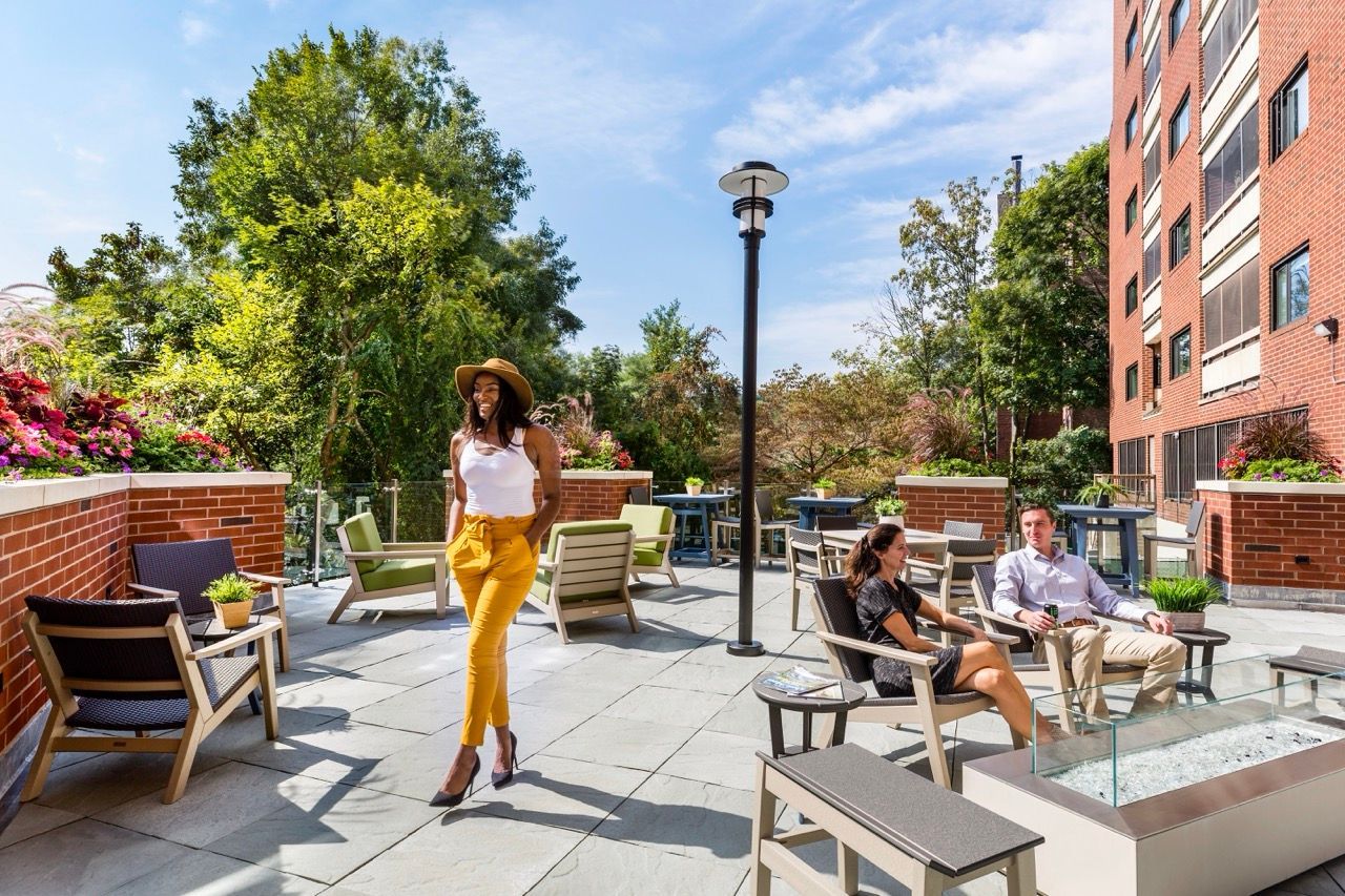 A woman walking on a sunny outdoor rooftop lounge with seating and greenery.