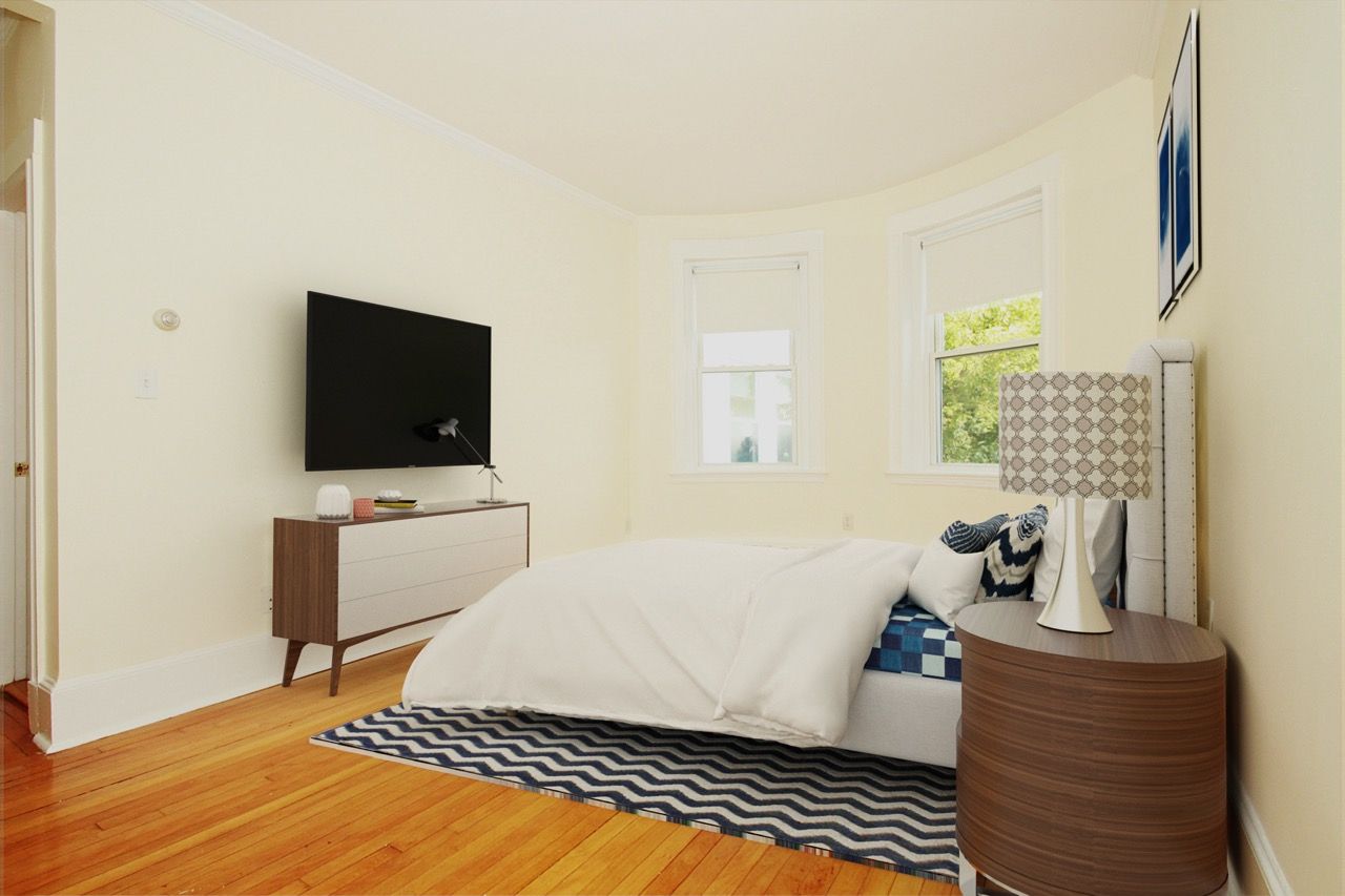 Bright apartment bedroom with a bed, dresser, nightstand, wall-mounted TV, and two windows.
