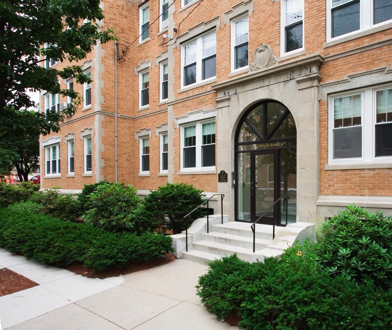 Brick apartment building entrance with an arched glass door, concrete steps, and trimmed shrubs.