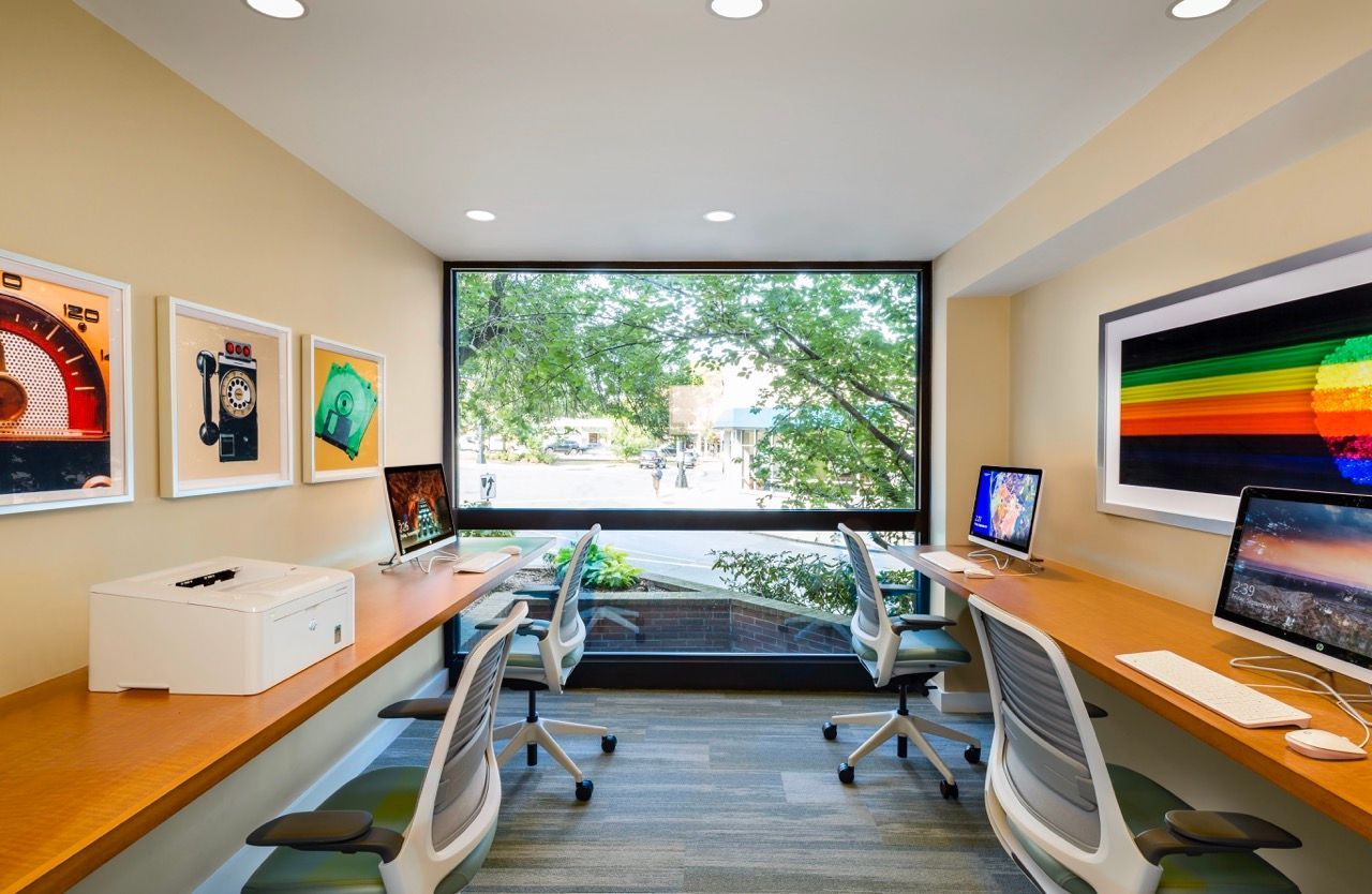 Community office area with long desks, ergonomic chairs, and a large window overlooking trees.