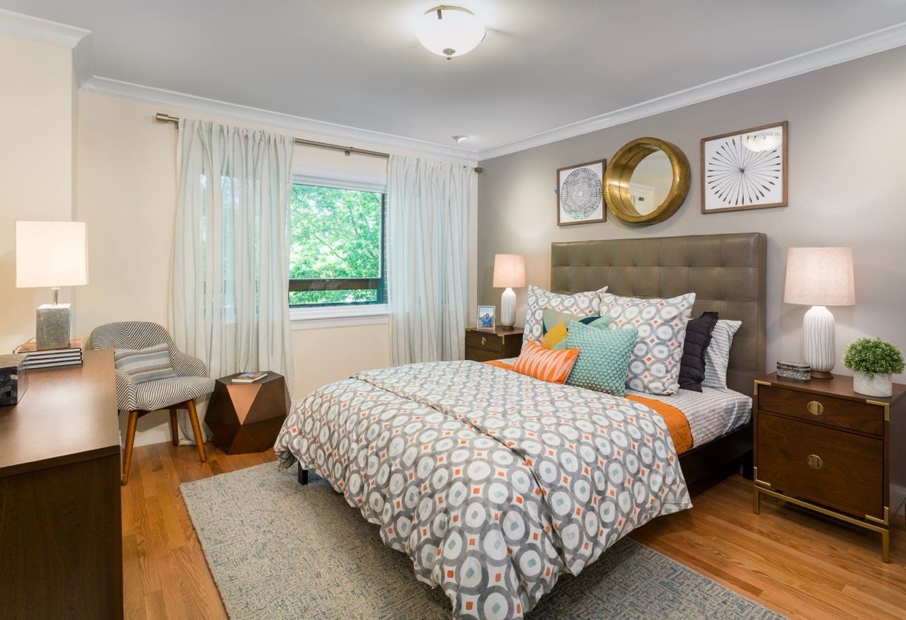 Bedroom in a modern apartment with a patterned quilt, nightstands, and a window with sheer curtains.