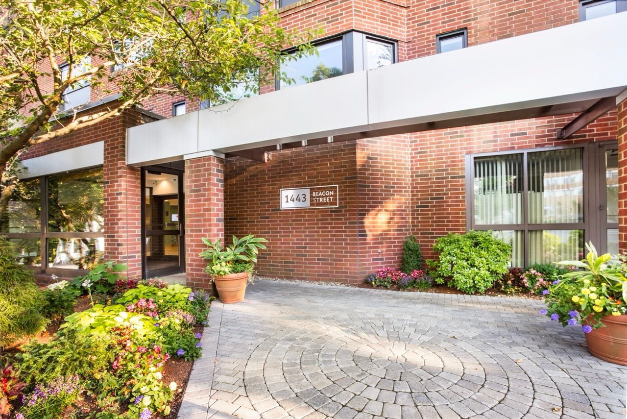 Brick apartment building entrance with a covered walkway and landscaped planters along the path.