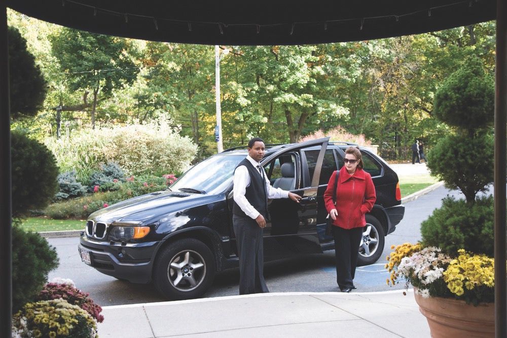 Two adults stand by a dark SUV with the driver's door open at a landscaped apartment entrance.