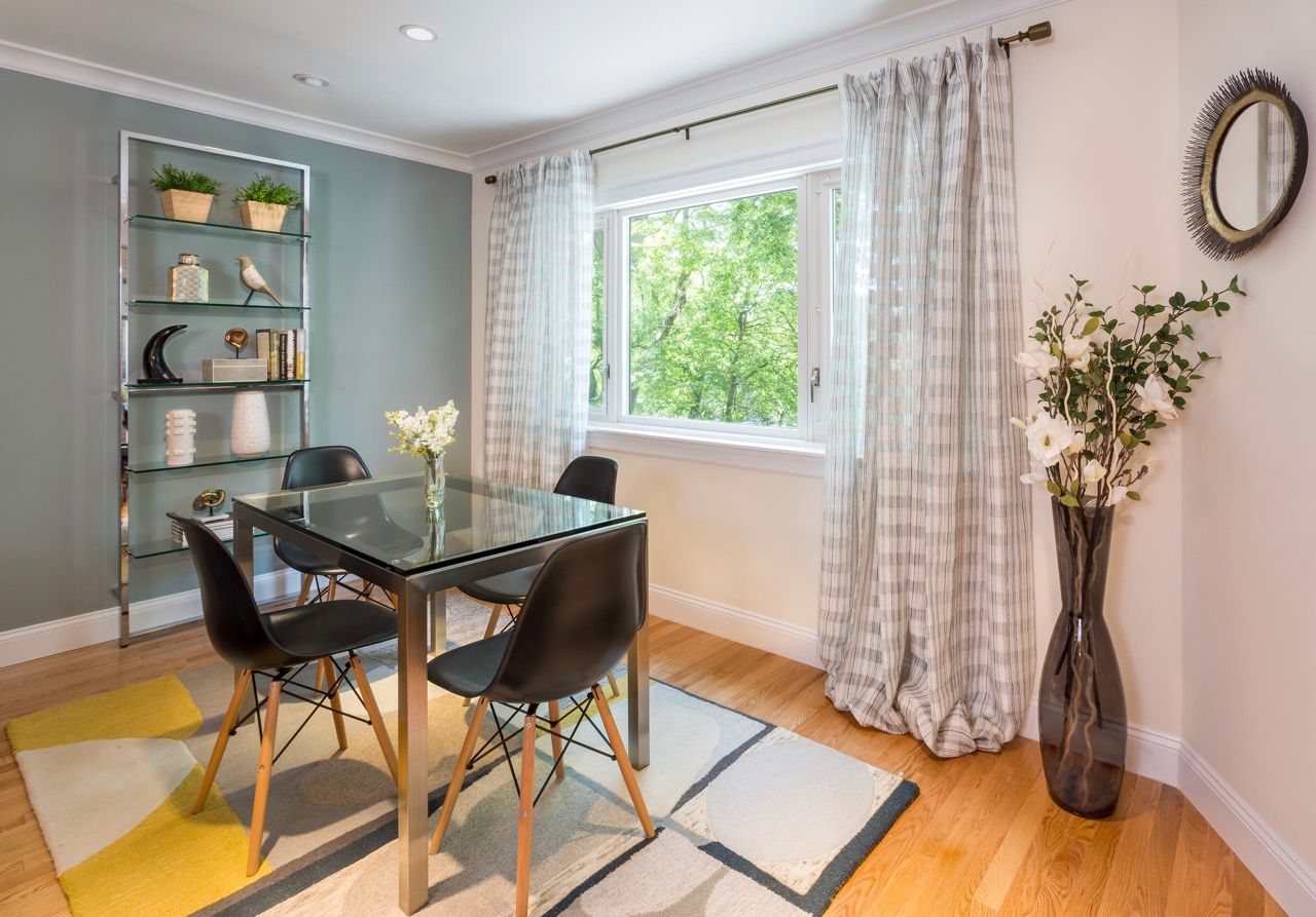 Dining area with glass-top table, four black chairs, glass display shelf, and a large window with striped curtains.