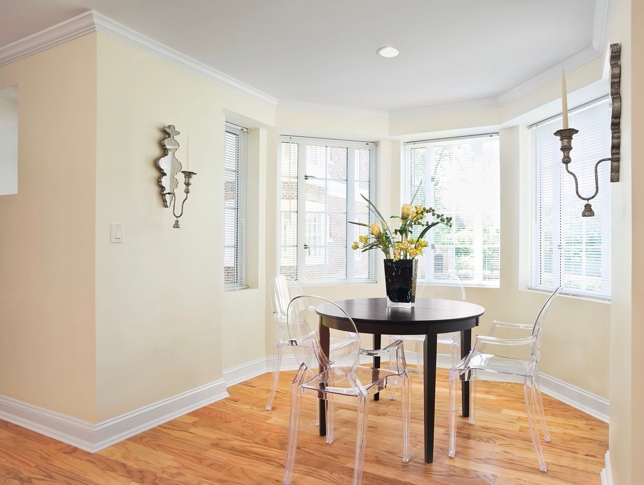 Dining area inside an apartment featuring a round table, clear acrylic chairs, and bay windows.