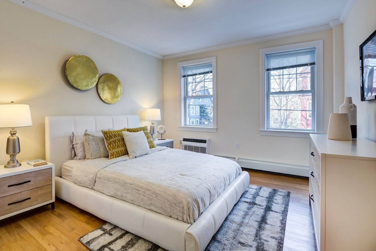 Bright modern apartment bedroom with a white upholstered bed, two nightstands, and a dresser.