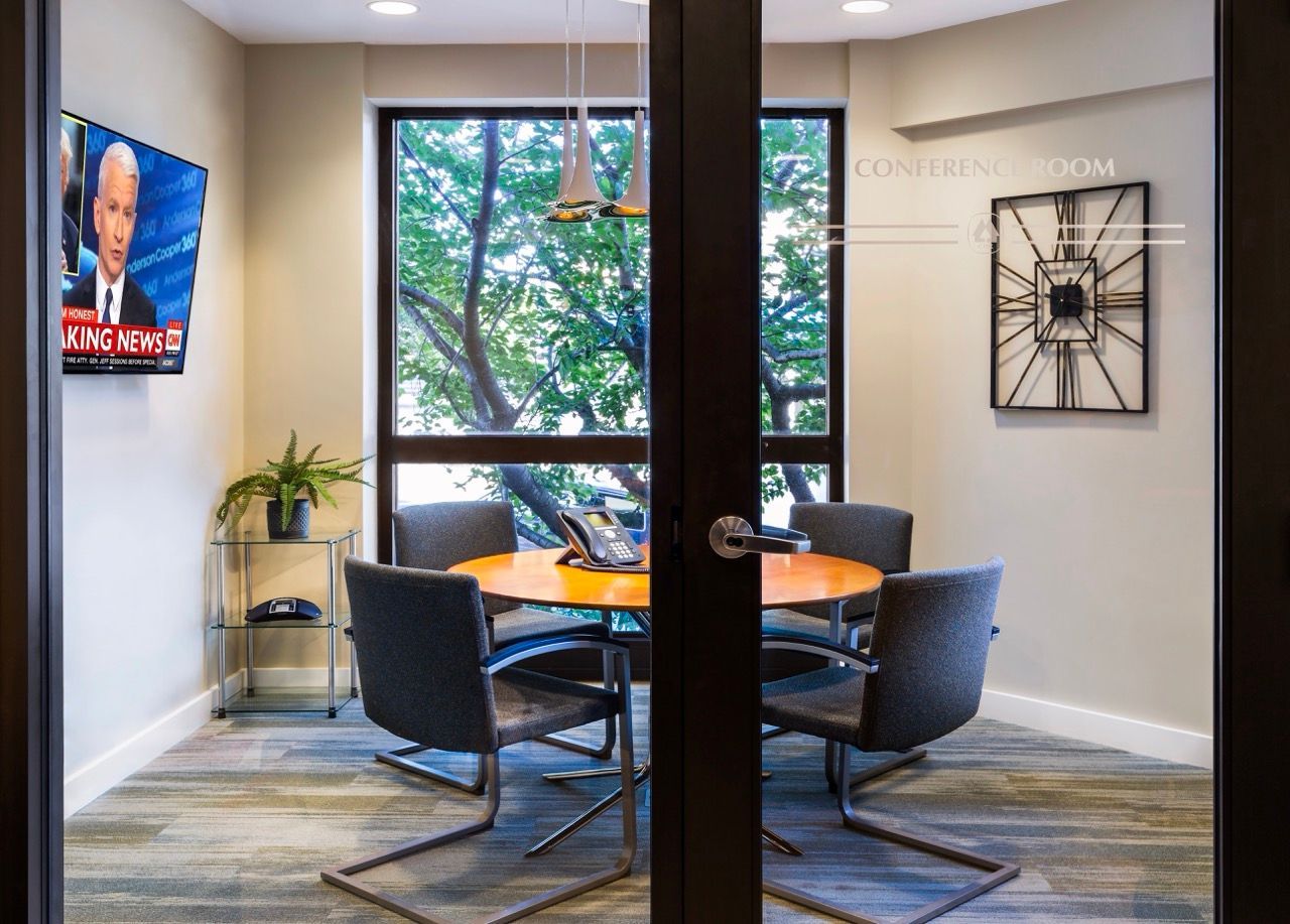 Conference room with a round wooden table, four gray chairs, glass doors, and a window view.