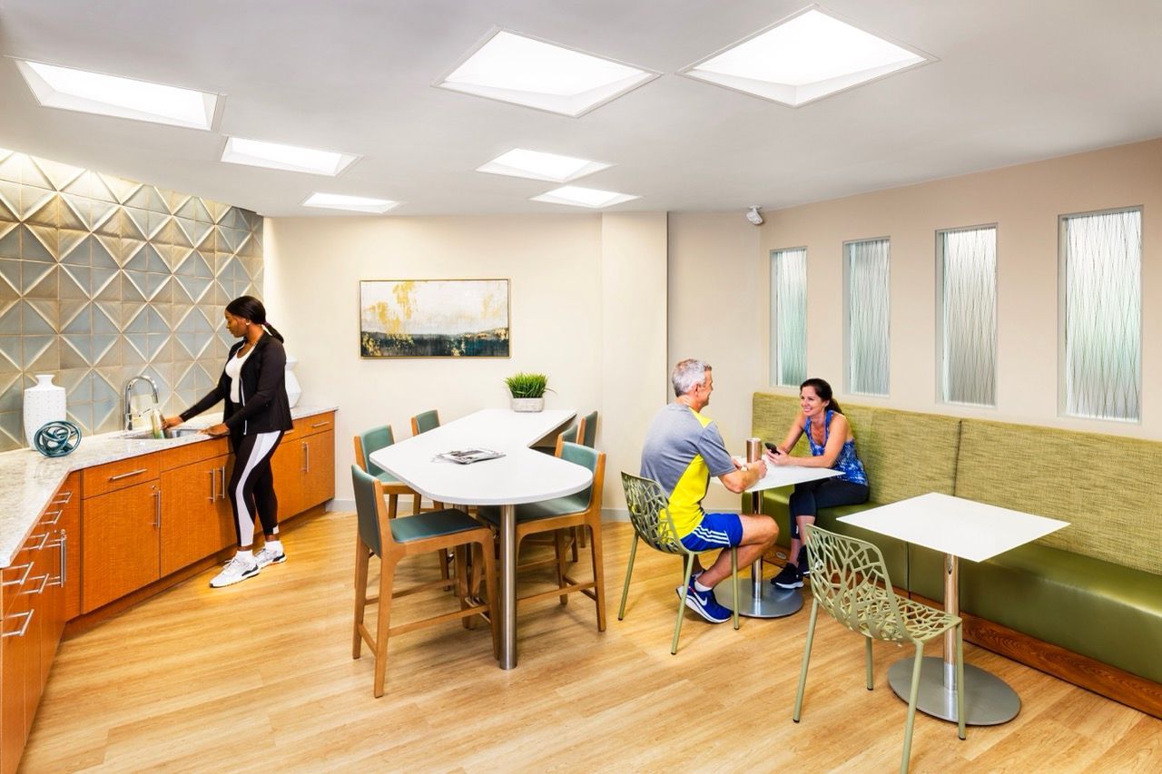 Two people chat at a white table in a bright communal lounge with a kitchenette and seating.