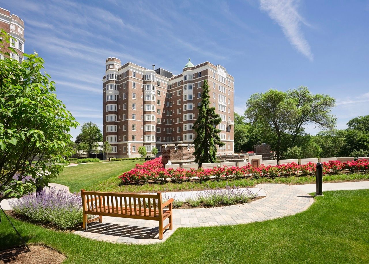 Exterior view of a brick apartment building with a manicured lawn, flower beds, and a wooden bench.