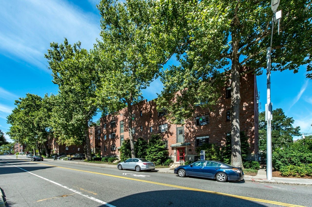 Exterior brick apartment building on a tree-lined street with parked cars.