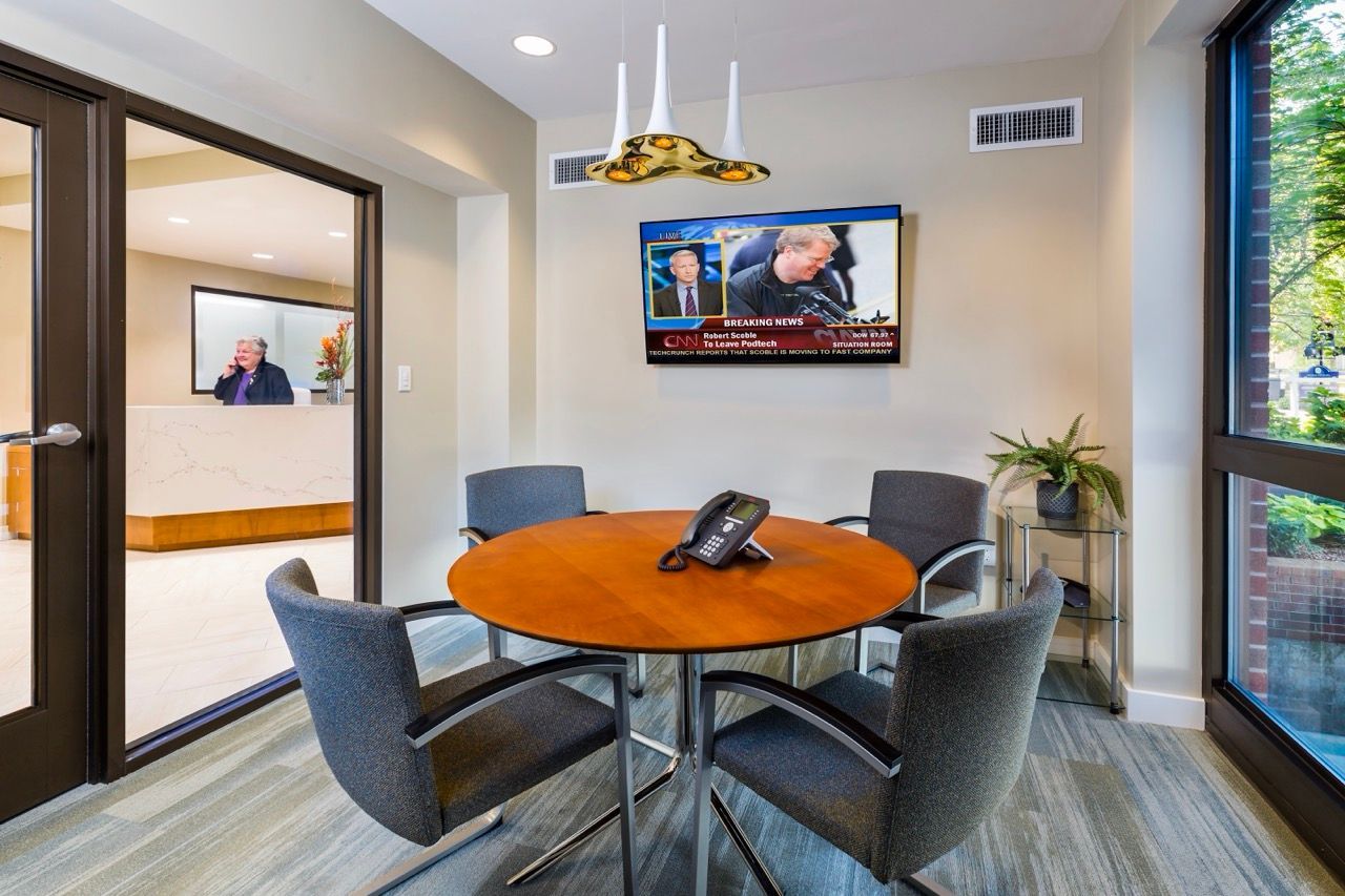 Modern meeting room with round wooden table, four gray chairs, wall TV, and glass entry.