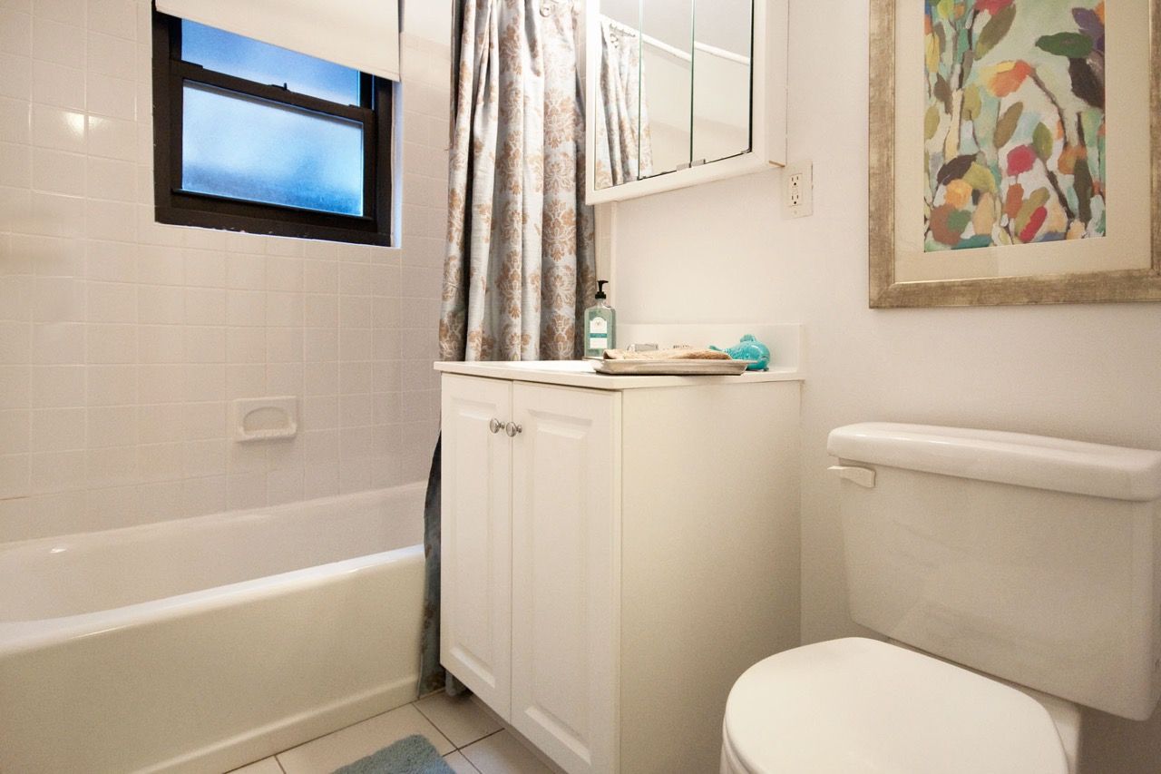 Small white bathroom with tub, vanity cabinet, and toilet; window above tub and patterned shower curtain.