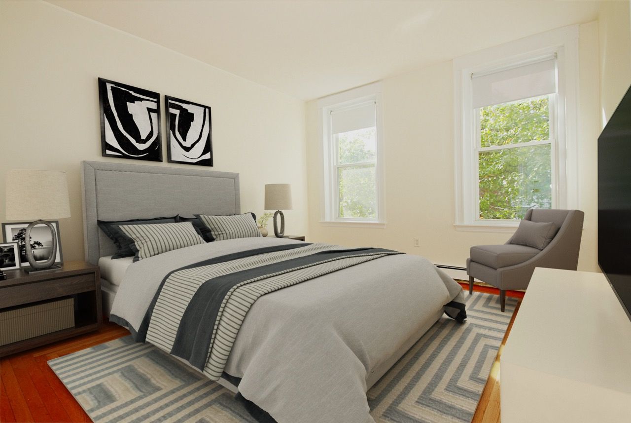 Bedroom in an apartment with a gray upholstered bed, two nightstands, lamps, and a chair by the windows.