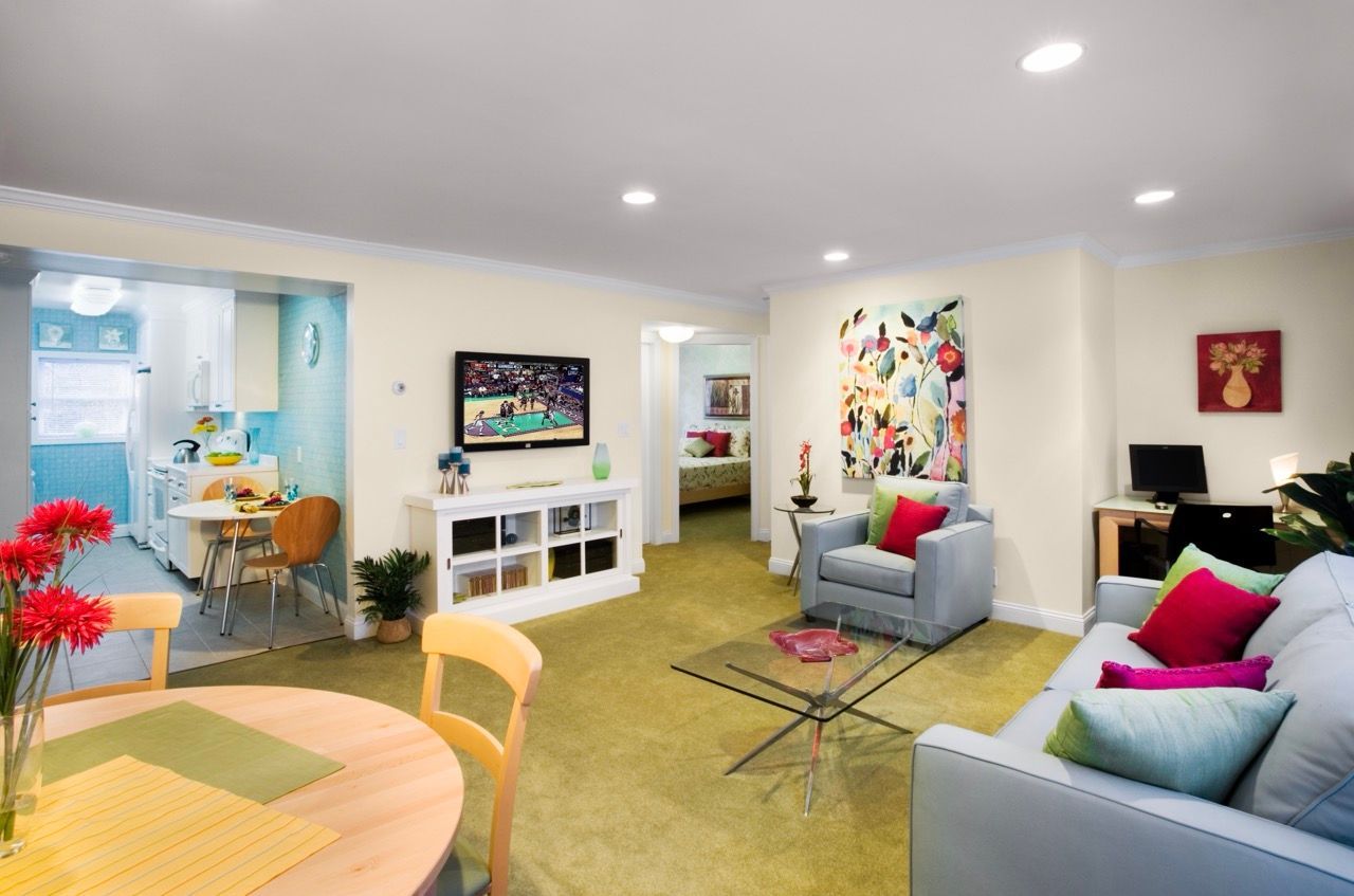 Bright living room in an apartment with pastel walls, light furniture, and an open kitchen visible in the background.