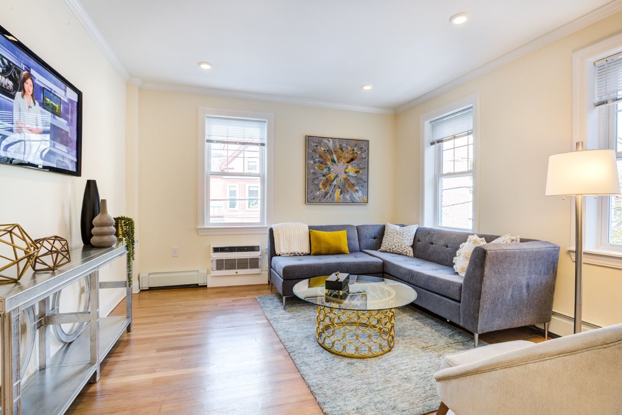 Bright living room in a modern apartment with a gray sectional sofa, glass coffee table, and large windows.